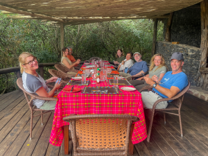 Eight people seated around a long outdoor table with red plaid cloth on a covered wooden deck.