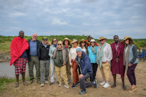 Group of fourteen people outdoors near water, two in Maasai attire, others in casual and outdoor clothing.