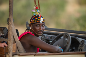 Man in colorful traditional beaded attire and headpiece seated in an open vehicle driver's seat.