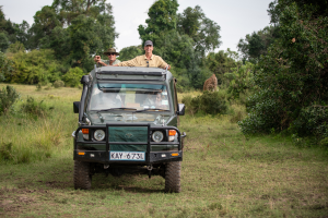 Safari vehicle with people parked on grassy path near trees and giraffe in background