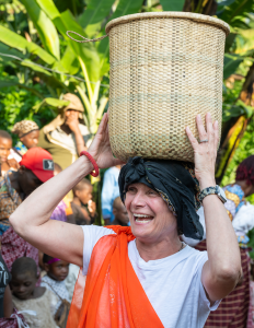 Woman in white shirt and orange garment balancing large woven basket on head outdoors.