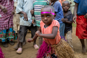 Child wearing pink headband and grass skirt dancing outdoors with other children around.