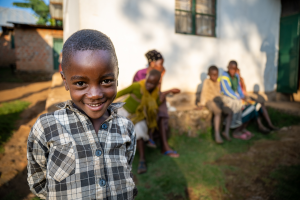 Young boy in a checkered shirt smiling outdoors with children and an adult sitting nearby.