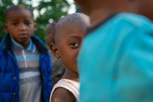 Close-up of a young boy partially hidden by another person outdoors with children blurred behind.
