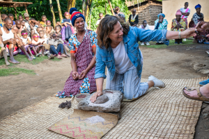 Woman in denim jacket grinding grain on stone while others watch outdoors in a rural setting