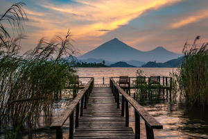 Wooden dock with chairs extends into lake framed by tall grasses and mountains at sunset.