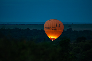 Hot air balloon with SKYSHIP CO. illuminated at burner in a grassy, forested landscape at dusk