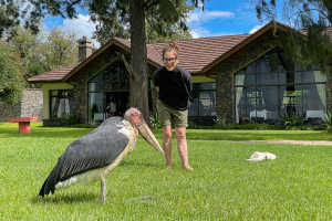 Person in black shirt and green shorts standing on grass near a large bird in front of a stone building.
