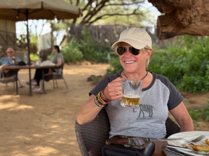 Woman wearing sunglasses, white cap, and bracelets, holding a beer mug at an outdoor table.