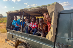 Children sitting and waving from the back of a green covered open truck on a dirt road.