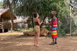 A woman in shorts and tank top talks to a person in colorful traditional attire outdoors.