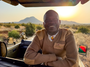 Man wearing a brown sweater with a beaded necklace leaning on a vehicle window at sunset.