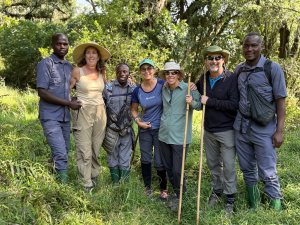 Seven people in outdoor clothing and uniforms pose with walking sticks in a grassy wooded area.