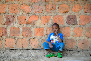 Boy in blue shirt and rolled-up pants sitting on the ground against a brick wall.