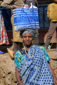 Woman in patterned clothing balancing a blue and white woven basket on her head outdoors