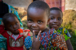 Three children outdoors, one smiling and making a peace sign wearing colorful patterned clothing.