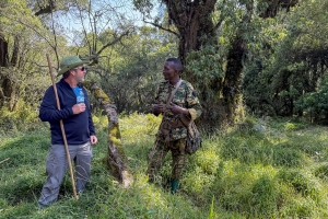 Two men talking in a dense forest, one in outdoor gear with a walking stick, the other in camouflage.