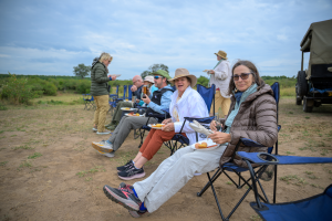 People seated in outdoor folding chairs eating food with a vehicle parked nearby and greenery background.