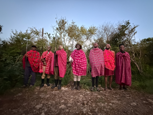 Seven people in red plaid traditional clothing stand on a dirt path with spears and greenery behind them.