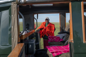 Man wearing red and orange plaid garment stands outside safari vehicle on grassy plain.