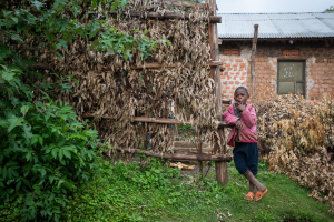 Boy in pink jacket and orange sandals leaning on a wooden structure with dried leaves near a brick house.