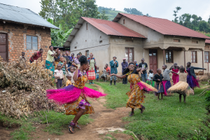 Two girls in colorful traditional dresses dance outdoors while a group watches and claps.