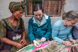 Three women weaving colorful straw mats while seated in front of a wall with a window.