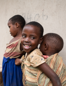 Three children stand against a wall, one carrying a younger child on their back with a cloth wrap.