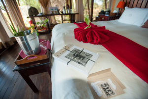 Bed with white sheets and red runner, elephant-designed paper, metal bucket with bottle and greenery on table.