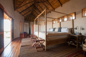 Bedroom with wooden four-poster bed, thatched ceiling, wooden floors, luggage racks, and sliding door.