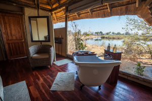 Bathroom with wooden floor, white freestanding tub, wicker chair, and river view through screened windows.