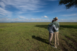 Couple embraces while looking over expansive grassy plain under a partly cloudy sky.