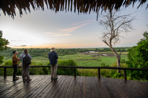 Three people stand on a wooden deck under a thatched roof overlooking a savanna with trees and a riverbed at sunset