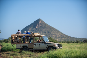 People on an open-sided safari vehicle in grassy terrain with a pyramid-shaped hill in the background.