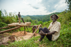 Three men woodworking logs outdoors in a green hilly rural landscape with dense vegetation.