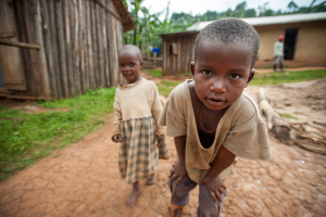 Two young barefoot children wearing worn clothes stand on a dirt path near wooden buildings.