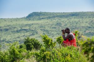 Person in red traditional clothing using binoculars on a grassy hill with green hills in background