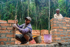 Two men building brick walls outdoors with construction tools and forest background