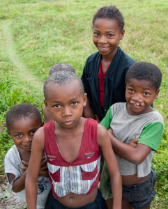 Five children standing on grass, four facing the camera with varied expressions, one partially hidden.