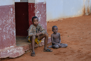 Two boys sitting on the ground outside a red and white painted building on reddish soil.