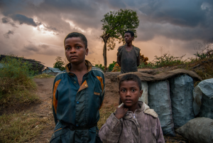 Two boys stand near large covered sacks outdoors with a woman and cloudy dusk sky behind them