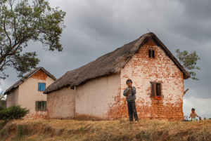 Two children stand near brick houses with thatched roofs in a rural area under cloudy sky