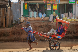 Man pulls rickshaw with passenger past a roadside store with sacks and bicycles.
