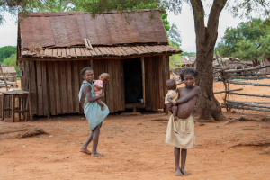 Two barefoot children each holding a baby stand in a rural area with wooden houses and red soil.
