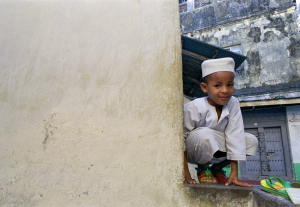 Young boy in traditional white clothing and cap crouching by a beige wall near colorful sandals.