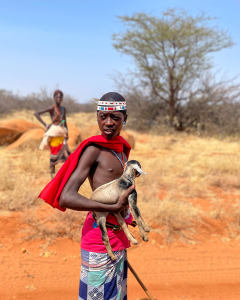 Young person in traditional attire holds a small goat in a dry, grassy landscape with a tree.