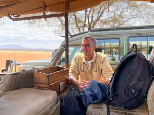 Man wearing glasses and beige shirt sitting in open-top safari vehicle with backpacks and jacket.