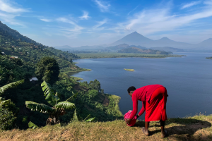 Person in red near grassy hill edge overlooking lake, hills, and distant mountains under blue sky.